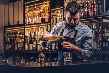 Stylish brutal barman in a shirt and apron makes a cocktail at bar counter background.