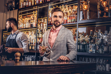 Stylish handsome male in an elegant suit holds a glass of whiskey at bar counter background.