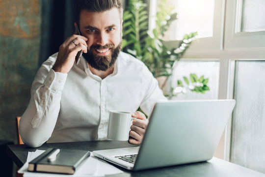 Young smiling bearded businessman is sitting at table in front of laptop, drinking coffee, talking on cell phone. Telephone conversations, remote work, online training, education, marketing, working.