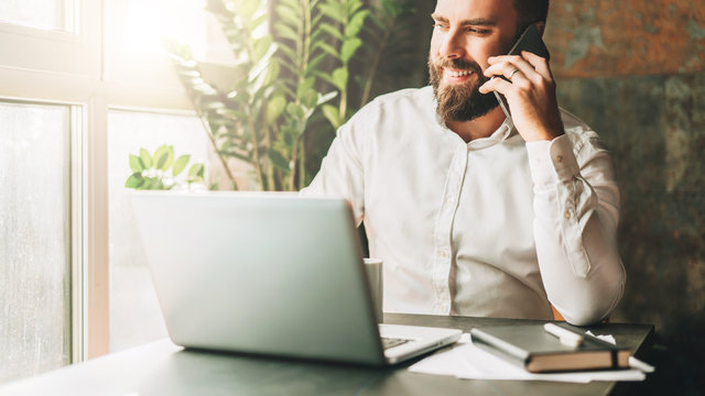 Young smiling bearded businessman is sitting at table in front of laptop, drinking coffee, talking on cell phone. Telephone conversations, distance work, online marketing, education, e-commerce.