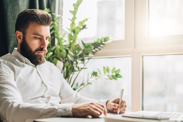 Young bearded businessman is sitting at table in office in front of laptop near window and writing in notebook. Man takes notes in a notepad. Distance work, online marketing, education, e-learning.