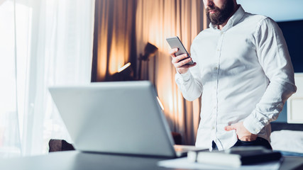Young serious bearded businessman standing near table in front of laptop, using smartphone. Man checks e-mail, blogging, planning. Online marketing, education, e-learning, social network, e-commerce.