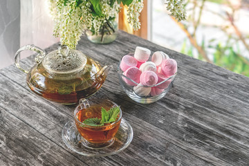 Mint tea in glass Cup on the background of a vase with marshmallows and a transparent glass teapot in a Sunny summer morning.