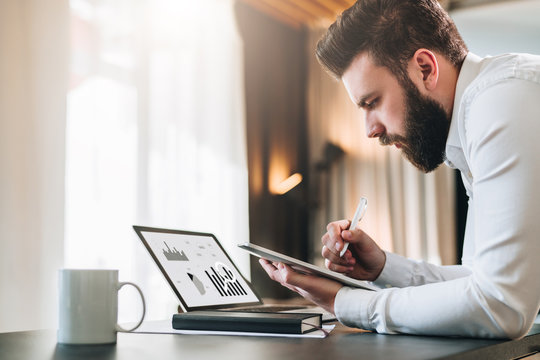 Young bearded businessman sitting at desk in front of laptop with graphis, charts, diagrams on screen, making notes in digital tablet. Freelancer works. Student learning online. Online marketing.