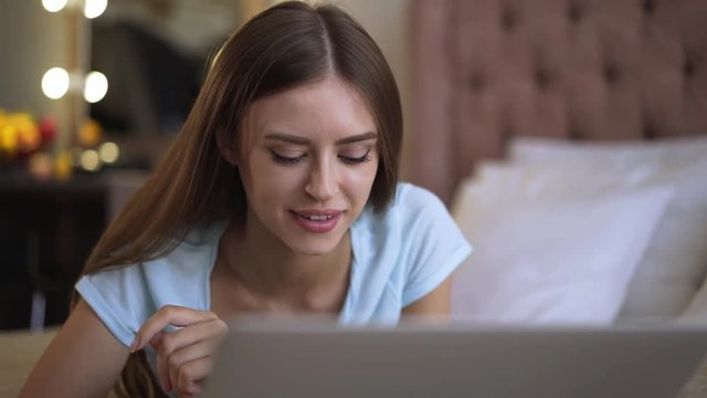 Portrait of a smling young woman with long fair hair lying on her bed and web surfing on her laptop computer. Slider real time medium shot