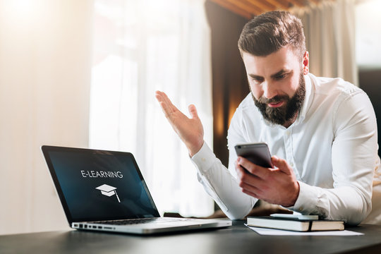 Young bearded businessman in a white shirt is sitting at a table in front of a laptop with an incription e-learning on screen and is happily looking at screen of smartphone, raising his hands up.