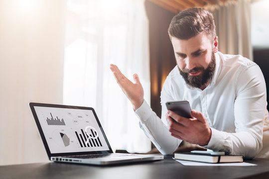 Young bearded businessman in white shirt is sitting at table in front of laptop with graphs, charts, diagrams on screen and is happily looking at screen of smartphone, raising his hands up.