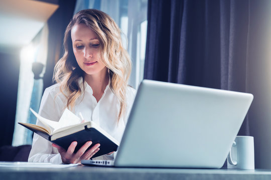 Young businesswoman in shirt is sits in office at table in front of computer and reads notes in notebook.Student does homework, girl reads book. Online marketing, education, e-learning, distance work.