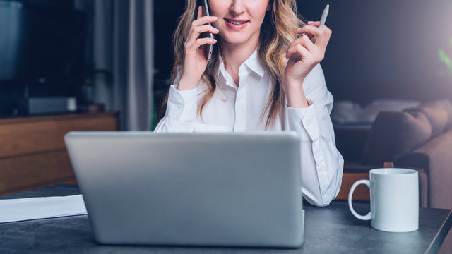 Young businesswoman in shirt is sitting in office at table in front of computer, talking on cell phone, holding pen in her hand. Telephone conversation, online marketing, education, e-learning.