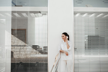 Portrait of laughing female person standing on balcony and using gadget for communication. Office blinds on background. Copy space in left side