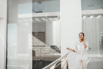 Portrait of female person conversing on phone outdoors and laughing. Office louvers on background. Copy space in left side