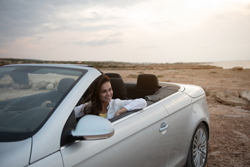 Pleasant time. Positive young charming woman is sitting in luxury cabriolet while looking aside with smile. She is enjoying summer trip abroad. Active lifestyle concept
