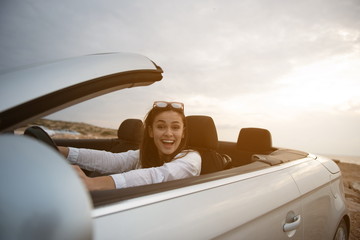 Unexpected thing. Portrait of surprised young positive female is looking at camera with wonder while sitting in fashionable modern car with open roof. Travel abroad and active lifestyle concept