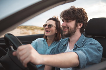 Fototapeta premium Summertime. Cheerful young pleasant male and female are travelling by luxury car. Focus on cute bearded boyfriend is holding on to steering wheel and expressing gladness. Romantic trip concept