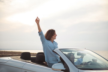 Full of joy. Cheerful young female friends are travelling by car. They are enjoying their summer vacation. Girl with raised hand is standing in cabriolet with open roof expressing happiness