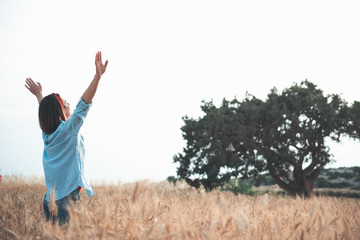 Freedom of mind concept. Back view of gorgeous girl is standing in golden wheat field with raised hands and expressing happiness. Copy space in the right side