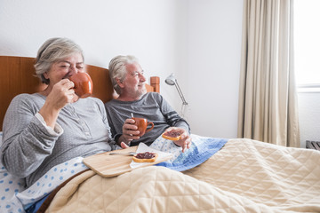 nice happy morning at home doing breakfast at bed with a lot of smiles and laugh for adult senrio caucasian couple married and in relationship. daily retired people scene in the bedroom for happiness