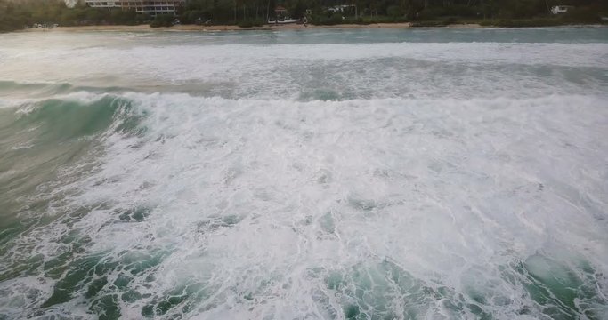 Drone flying backwards from peaceful ocean coastline revealing big foaming waves washing the shore on a sunny summer day