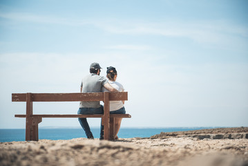 Nice time. Full length back view of relaxed couple is sitting on the bench in front of sea with colorful blue sky in background and enjoying great view. Man is hugging his girlfriend. Copy space