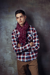 Portrait of young trendy handsome man with short dark hair wearing checkered shirt and brown trousers standing and posing against gray concrete wall