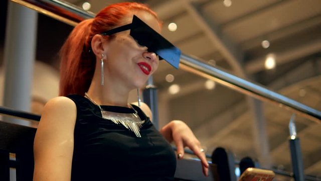 Stylish fashion red hair woman in black dress and square glasses watches mobile phone news