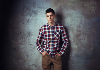 Portrait of young trendy handsome man with short dark hair wearing checkered shirt and brown trousers standing and posing against gray concrete wall