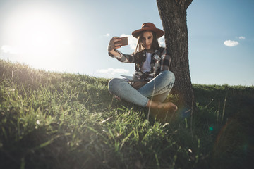 Selfie time. Full length of stylish girl taking picture of herself while sitting on grass. She is posing at camera with playful countenance. Copy space in left side