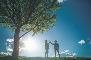 Full length of young couple walking in bright sunny day. They are standing by nice green tree opposite to each other and gently touching their hands