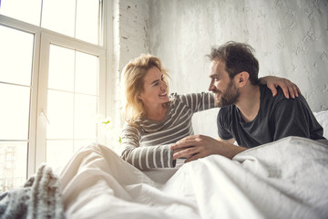 Happy two smiling people who are in love. They are meeting morning together sitting in bed and showing fondness to each other. Lady is putting arm around partner neck and looking at him with love