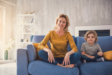 Smiling parent is sitting with her delighted kid on sofa. They are happily spending time together in cozy apartment and looking at camera. Mom and son together concept