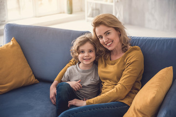 Cheerful family are relaxing together in sitting-room. Mom is embracing smiling kid while they are looking at camera with joy. Motherhood concept