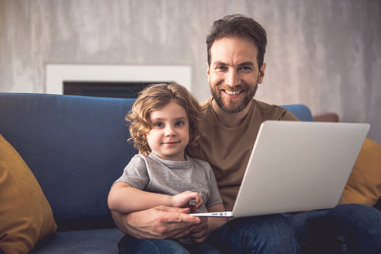 Happy father and son are sitting on sofa with laptop. Parent is holding gadget on knees while hugging his child. They are grinning and looking at camera