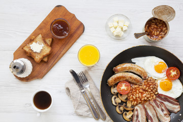 Full English breakfast in a pan with fried eggs, bacon, beans, sausages  and toasts on white wooden background, top view. From above, flat lay.