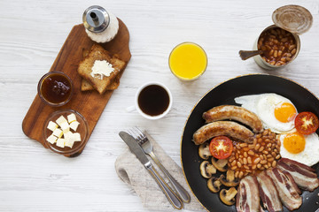 Top view, full English breakfast in a pan with fried eggs, beans, sausages, bacon and toasts on white wooden background. Overhead shot, flat lay.