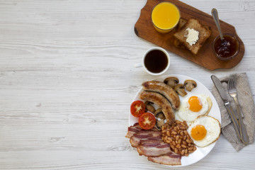 Flat lay, full english breakfast with fried eggs, sausages, bacon, beans and toasts. White wooden background. Top view. Copy space.