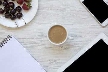 Work space with smartphone, strawberries, cherries, notepad and latte in the center on white wooden background. Top view.