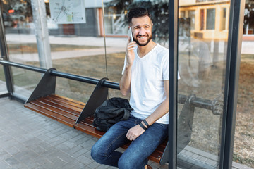 photos of a beautiful stylish Guy, in a blank white t-shirt, a hipster talking on the phone, and sitting at the bus stop waiting for transport, can be used for advertising, text insertion