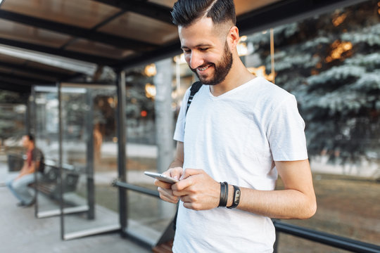 photo of a beautiful stylish Guy, in a blank white t-shirt, hipster looking at the phone standing at the bus stop waiting for transport, can be used for advertising, text insertion