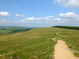 a long winding footpath running into the distance in stoodley moor in yorkshire with fields and farms in the distance with the pennine hills on the horizon on a sunny day with blue sky and clouds