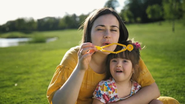 Mother sitting on green grass meadow with sweet little toddler daughter with down syndrome and blowing soap bubbles together. Happy family spending leisure together in summer nature