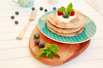 Turquoise plate with white dots with stack of pancakes, wooden plate, blue serviette underneath, sour cream topping with raspberries, blueberries and fresh mint, fork, blue mug and glass with milk