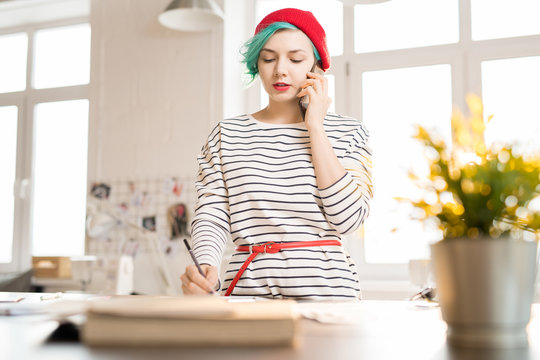 Waist up portrait of contemporary female fashion designer speaking by phone and taking notes while standing at work table in sunlit atelier workshop - Powered by Adobe