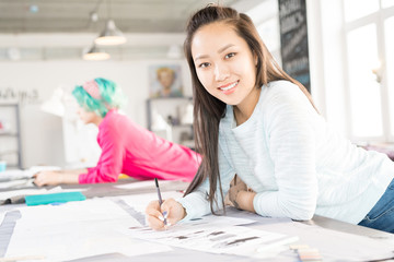 Portrait of smiling Asian woman looking at camera while drawing sketches at tailors table in modern sunlit atelier