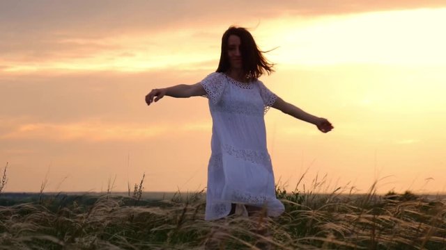 Beautiful girl wearing white dress running through beautiful field at sunset. Young woman jogging at the meadow and enjoying freedom. Summer leisure at nature concept.