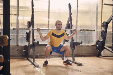 one young smiling man, wearing sport clothes, squat exercise with bar, in old beaten up gym interior. full lenght shot.