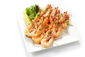 Roasted big black tiger shrimps skewers with herbs, piece of lemons and fresh vegetables on white square plate, isolated on white background with shadow, High angle top view, Focus at middle of food.