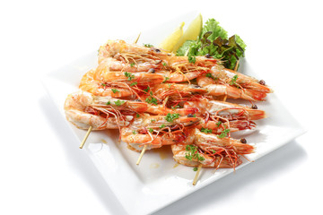 Grilled big black tiger shrimps skewers by bamboo sticks with herbs, piece of lemons & fresh vegetables on white square plate, isolated on white background with shadow, high angle view.