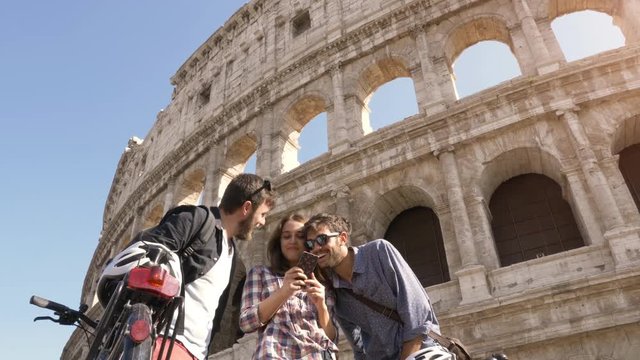 Three happy young friends tourists with bikes and backpacks at Colosseum in Rome using smartphones browsing selfies pictures on sunny day slow motion steadycam ground shot