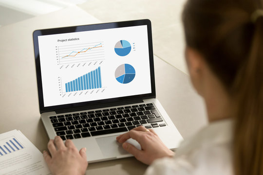 Young business woman in office working on laptop with project statistics on screen. Office worker reviewing successful positive business results. Corporate success concept. Close up, focus on screen.