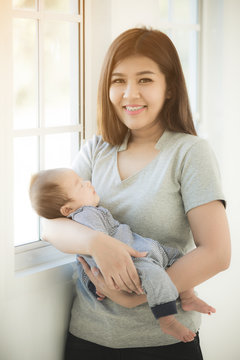Asian Mother Cuddling Baby Daughter At Home In Front Of Window